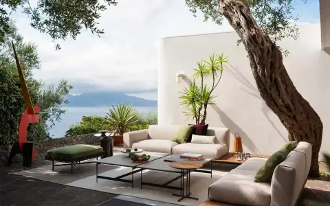 Arredare un terrazzo scoperto con divani chiari, tavolini bassi e un pouf verde, con vista mare e un ulivo secolare.