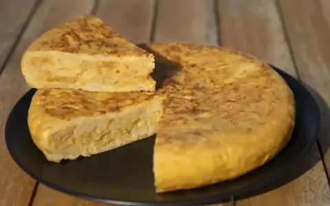 Una deliciosa tortilla de patatas, con una porción cortada y apilada, servida en un plato negro sobre una mesa de madera.