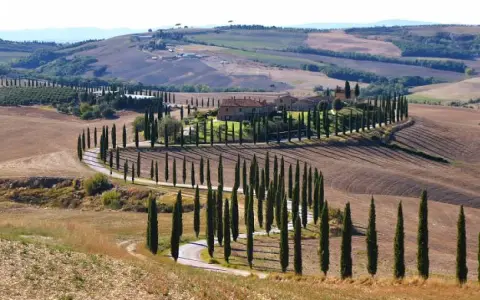 Strade sinuose e cipressi iconici nel parco della Val d'Orcia, con un casale in lontananza.
