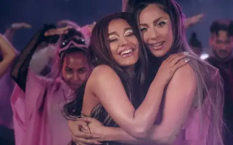 Ariana Grande i Lady Gaga przytulają się, otoczone tańczącymi ludźmi w różowych strojach. Czuć, że na nich "rain on me".