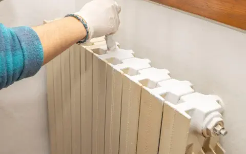 Mano con guanto bianco che dipinge un termosifone beige con vernice bianca, pronto per imbiancare la stanza.