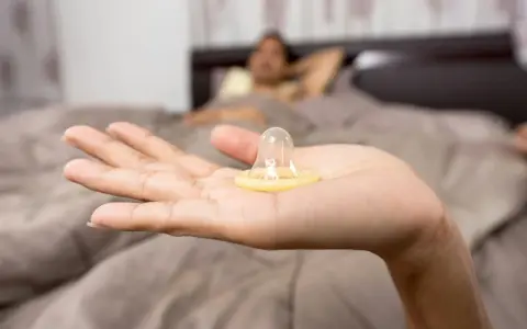 Mano sostiene un condón. En el fondo, una persona en la cama. La decisión de tener sexo oral sin condón es personal.
