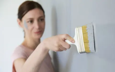 Une femme effectue une retouche peinture murale avec un pinceau gris. Une tache de peinture grise est visible sur sa joue.