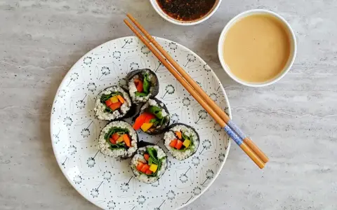 Sześć wegetariańskich rolek sushi z kolorowymi warzywami, przygotowanych z użyciem glonu do sushi, leży na talerzu obok sosów i pałeczek.