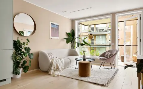 Jasne meble do salonu: puszysta sofa, okrągły stolik, aksamitny fotel i puf. Duże okno z widokiem na balkon.