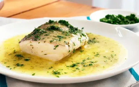 Un filete de merluza en salsa verde, con perejil picado, servido en un plato blanco.