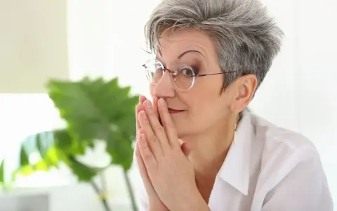 Elegancka kobieta z siwymi włosami w odmładzającej krótkiej fryzurze damskiej, w okularach, z dłońmi złożonymi przed twarzą.