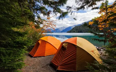 Två orangea tält står uppställda vid en sjö, omgivna av höstfärgad skog och berg. En perfekt plats för kamlunge camping.