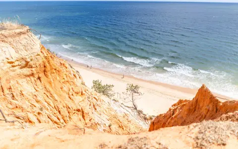 Orange cliffs overlook a sandy portugal strand algarve. Gentle waves lap the shore where a few people stroll.