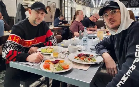 Dwóch mężczyzn, jeden w bluzie z napisem "Givenchy", drugi w bluzie "Supreme", siedzi przy stole w restauracji. Wyglądają jak bary raperzy, którzy wybrali się na wspólny posiłek.