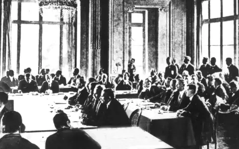 Männer in Anzügen sitzen an langen Tischen in einem prunkvollen Saal. Dies ist die Unterzeichnung des Versailler Vertrags, der den Ersten Weltkrieg beendete.