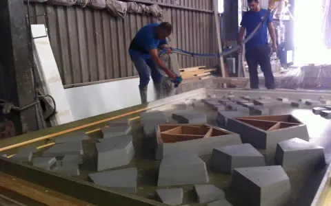 Dos operarios trabajan con material GRC en un taller, uno rocía con una manguera y el otro supervisa.