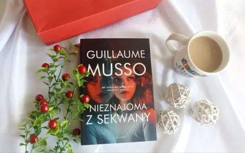 Książka "Nieznajoma z Sekwany" Guillaume'a Musso leży obok kubka kawy i czerwonych ozdobnych kulek.