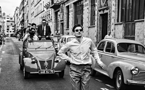 Un uomo con sigaretta in bocca, occhiali da sole e camicia a righe, in strada con auto d'epoca, evoca lo spirito de **la nouvelle vague**.