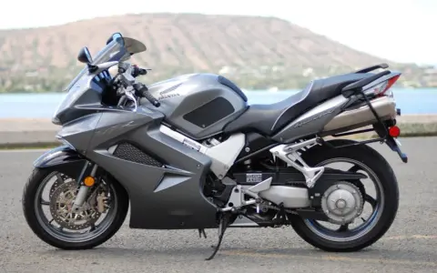 Szary motocykl Honda VFR 800 z widokiem na wodę i wzgórze w tle.