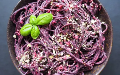 Fioletowy makaron spaghetti z buraczanym pesto, posypany orzechami i udekorowany listkiem bazylii.