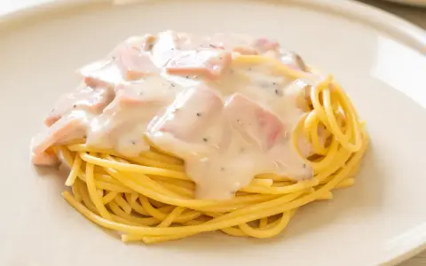 Spaghetti z kawałkami szynki w kremowym sosie śmietanowym, posypane pieprzem. Pyszne danie na talerzu.