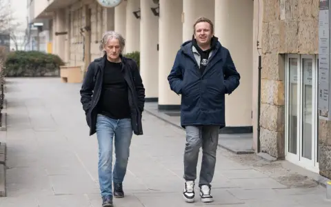 Jacek Podsiadło, ojciec Dawida, idzie ulicą w granatowej kurtce i jeansach. Obok niego młodszy mężczyzna w podobnym stroju.