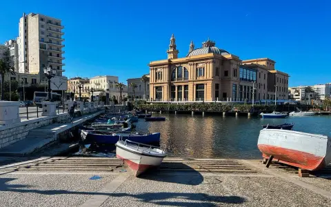 Barche ormeggiate nel porto di Bari, con il Varco della Vittoria e il Teatro Margherita sullo sfondo, sotto un cielo azzurro.