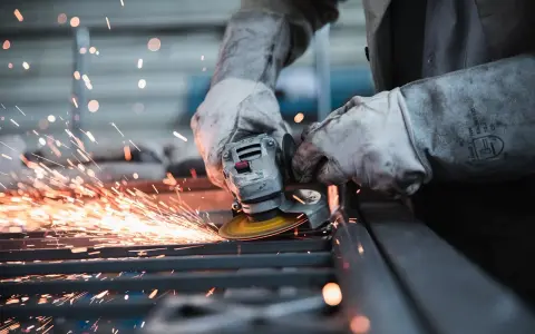 Pracownik w rękawicach szlifuje metal, sypią się iskry. Wypadek przy pracy? Protokół powypadkowy wymaga dokumentacji.