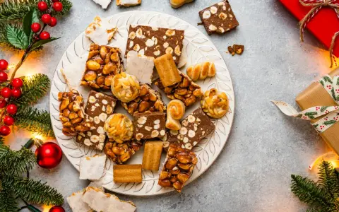 Plato con variedad de postres navideños, turrones, mazapanes y dulces de frutos secos, rodeado de ramas de abeto y luces.