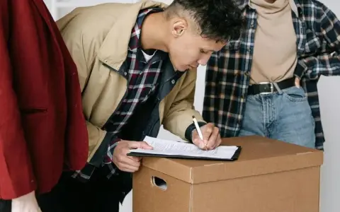 Joven firma contratos de alquiler sobre una caja de mudanza, acompañado de dos personas.