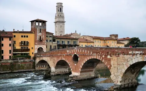 En vacker tegelbro korsar en flod i Verona, Italien. Färgglada hus och ett högt klocktorn syns i bakgrunden.