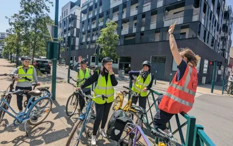 Des adultes souriants, casqués et en gilets fluo, prêts à apprendre à faire du vélo adulte. Ils sont avec leurs vélos, devant un immeuble moderne.