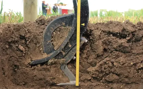 Czarne, metalowe elementy podeszwy płużnej zagłębiają się w ziemi. Obok miarka pokazuje głębokość orki.
