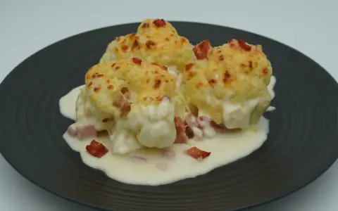 Tres porciones de coliflor gratinada con bechamel y trocitos de jamón, servidas en un plato oscuro.