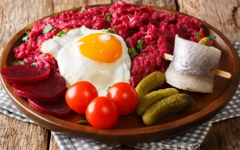 Herzhaftes Labskaus essen: Rote Bete, Spiegelei, Matjes, Cornichons und Tomaten auf einem Holzteller.