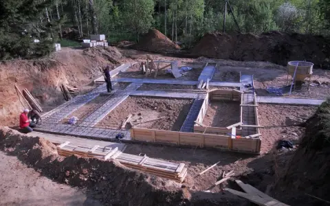 Budowa fundamentów domu. W wykopie widać zbrojenie i szalunki. Pracownicy przygotowują teren pod betonowanie. Nawet chudziak jest już gotowy.