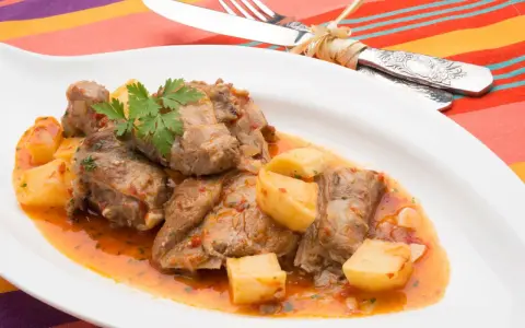 Sabroso cordero al chilindrón con patatas, servido en plato blanco con cubiertos atados con hilo.