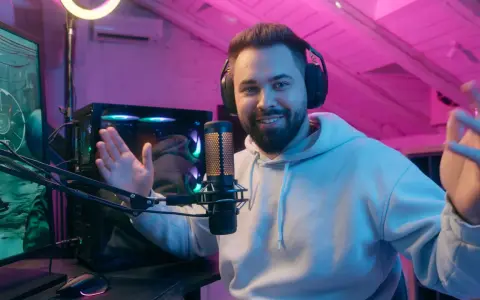 Uśmiechnięty streamer w słuchawkach, przy mikrofonie, tłumaczy, streaming co to jest. W tle komputer i różowe oświetlenie.