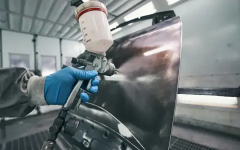 Ręka w niebieskiej rękawiczce trzyma pistolet lakierniczy, rozpylając farbę na element karoserii. Czas schnięcia szpachli na epoksyd jest kluczowy dla dalszych prac.