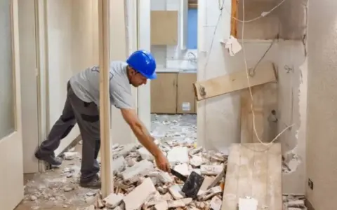 Hombre con casco azul rompiendo escombros con un mazo. Parece que él mismo **puedo tirar un tabique** para renovar la cocina.