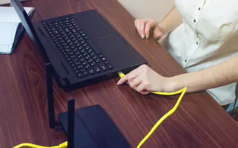 Ręka podłącza żółty kabel do laptopa, pokazując, czy można podłączyć monitor do laptopa. Obok leży router.