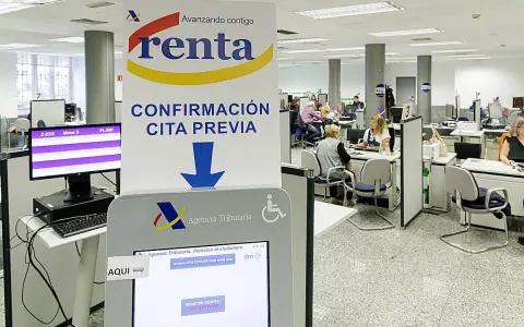 Oficina de la Agencia Tributaria con gente esperando. Un cartel indica "Confirmación cita previa" y una pantalla táctil para gestionar citas, resolviendo la duda de cuanto tarda Hacienda en cobrar la renta.