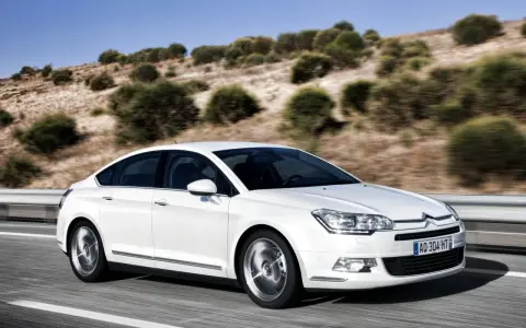 Biały Citroen C5 2.0 HDI pędzi po pustej drodze, otoczony suchymi, pagórkowatymi krajobrazami.