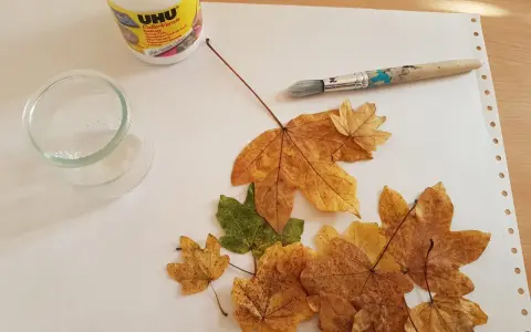Préparation d'une décoration avec des feuilles d'arbre séchées, de la colle UHU, un pinceau et un petit récipient en verre.
