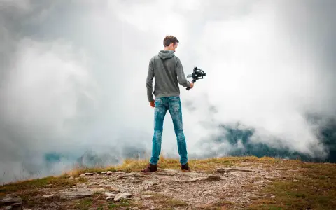 Joven en la cima de una montaña, sosteniendo un **gimbal** para estabilizar su cámara y capturar la vista entre las nubes.