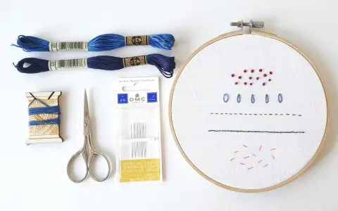Materiali per un tutorial ricamo: fili blu, forbici, aghi e tela con primi punti.