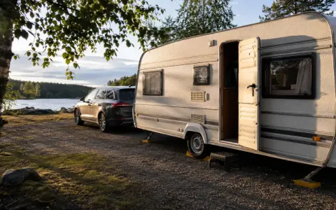 En husvagn och bil parkerad vid en sjö, redo för en husvagnssemester i Sverige. Solen lyser på den öppna dörren.