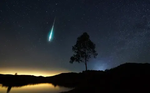 Un **obiettivo per fotografare le stelle** cattura una scia luminosa nel cielo notturno, con un albero solitario in silhouette e un lago calmo.