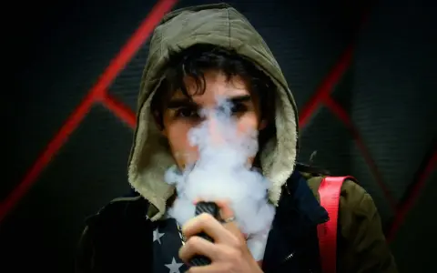 Joven con capucha exhala vapor de un vapeador, con un diseño de estrellas en su camiseta. Los nicokits son su secreto.