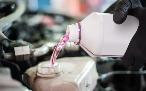 Mano con guante negro vierte líquido rosa, agua de motor coche, en depósito.
