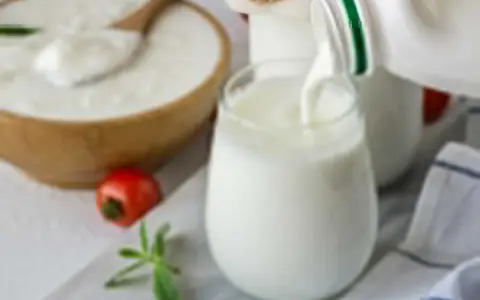 Ayran co to? Orzeźwiający napój mleczny, idealny na upalne dni, nalewany do szklanki. Obok miska z jogurtem i pomidorek.