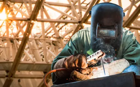 Soldador con máscara y guantes realiza soldadura con electrodo, creando chispas y humo bajo una estructura metálica iluminada por el sol.