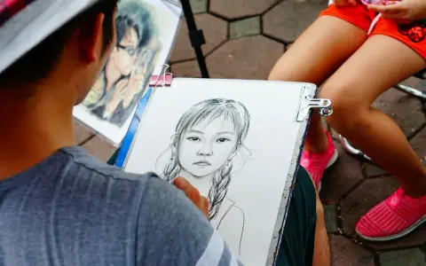 Artysta tworzy portret dziewczynki z warkoczami. Obok leży gotowy rysunek chłopca w okularach.