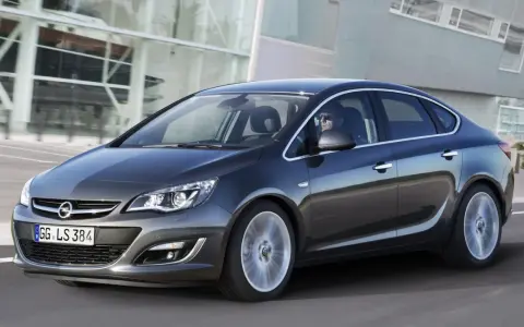 Ciemnoszary Opel Astra J 1.6 benzyna w ruchu, z kierowcą w okularach za kierownicą, na tle nowoczesnego budynku.